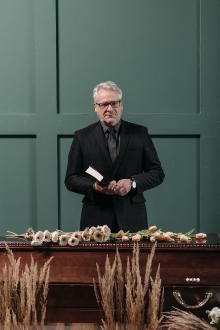A man with gray hair, glasses, and a somber expression stands behind a polished wooden casket, dressed in a black suit and tie, holding a small book or prayer card. In front of him, several white and 