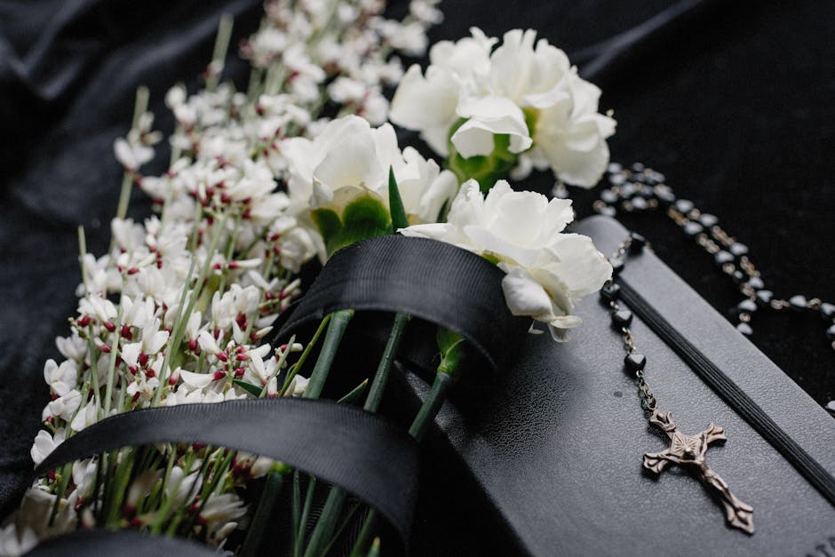 A close-up image of fresh white flowers, including chrysanthemums and small clustered blossoms, arranged with green stems, resting on a black surface. The flowers are partially wrapped with black ribb