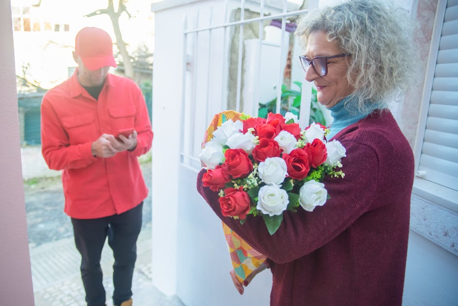 A woman with curly gray hair, wearing glasses, a blue turtleneck, and a burgundy sweater, holds a floral gift arrangement featuring red and white roses wrapped in colorful paper. She is standing near 