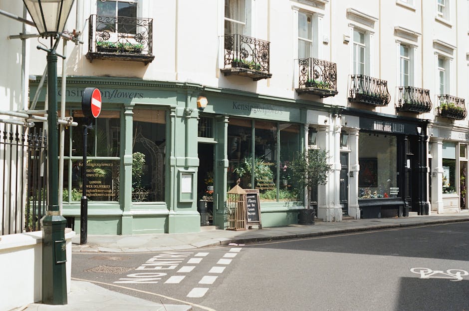 A charming flower shop with a pale green exterior and large display windows showcasing fresh floral arrangements. The shop has a black signboard above the entrance reading 'Kensington Flowers,' and a