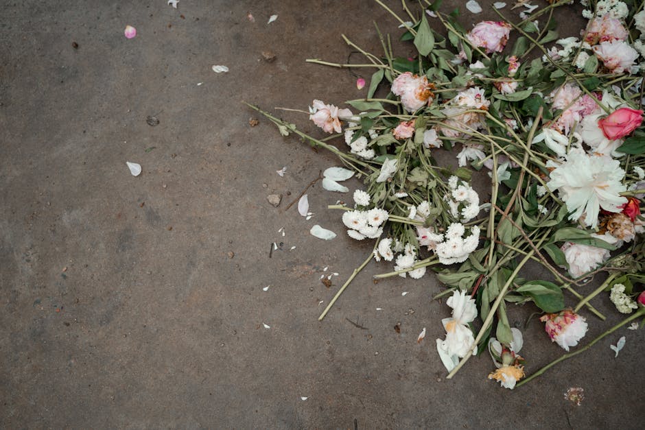 A pile of discarded floral waste on a concrete surface, consisting of various flowers including white chrysanthemums, pink roses, and white lilies, with green stems and leaves interspersed. Some petal