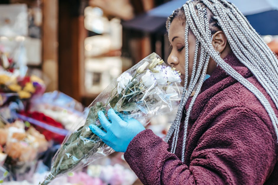 A woman with braided silver hair, wearing a burgundy fleece jacket and blue gloves, is holding a bouquet of white and pale green flowers wrapped in clear plastic at a florist shop. The bouquet feature