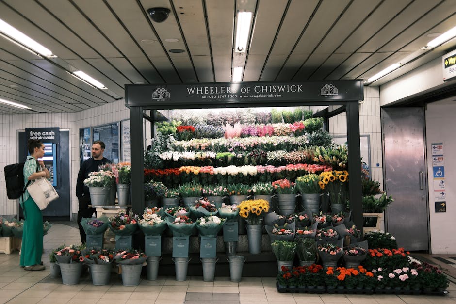 A floral display stand labeled 'Wheeler of Chiswick' in an underground station corridor, featuring an assortment of fresh flowers arranged in metallic buckets and wrapping paper. The bouquet arrangeme