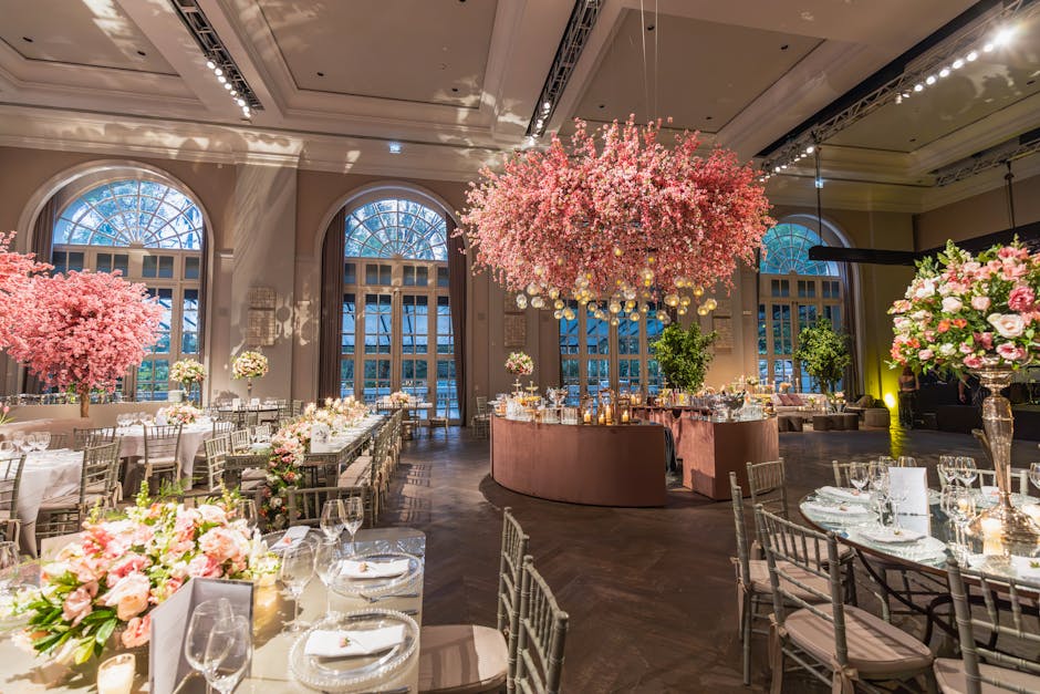 An elegant ballroom decorated for a wedding event with large, arched windows allowing natural light to illuminate the space. The central focus is a grand floral display featuring a massive, suspended