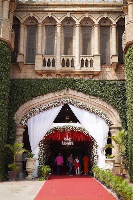 An elegant floral arrangement adorns the archway of a historic stone building, decorated for a wedding event. The arch is outlined with a lush garland of predominantly white and blush pink roses, mixe